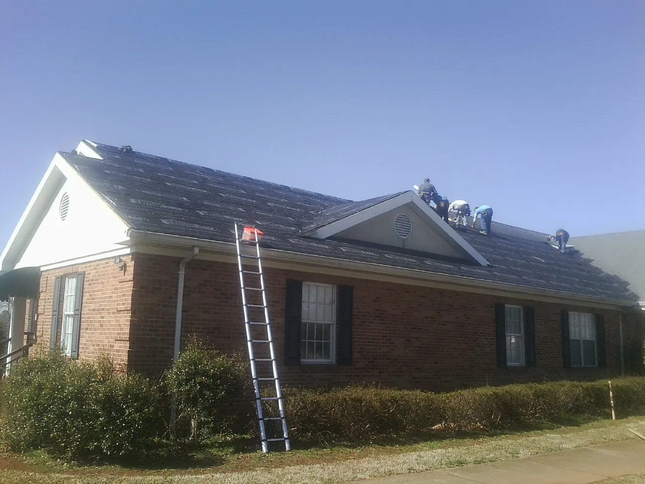 Crew tearing shingles off a brick ranch roof with a ladder set — Emergency Roof Repair work by our crew in Simms