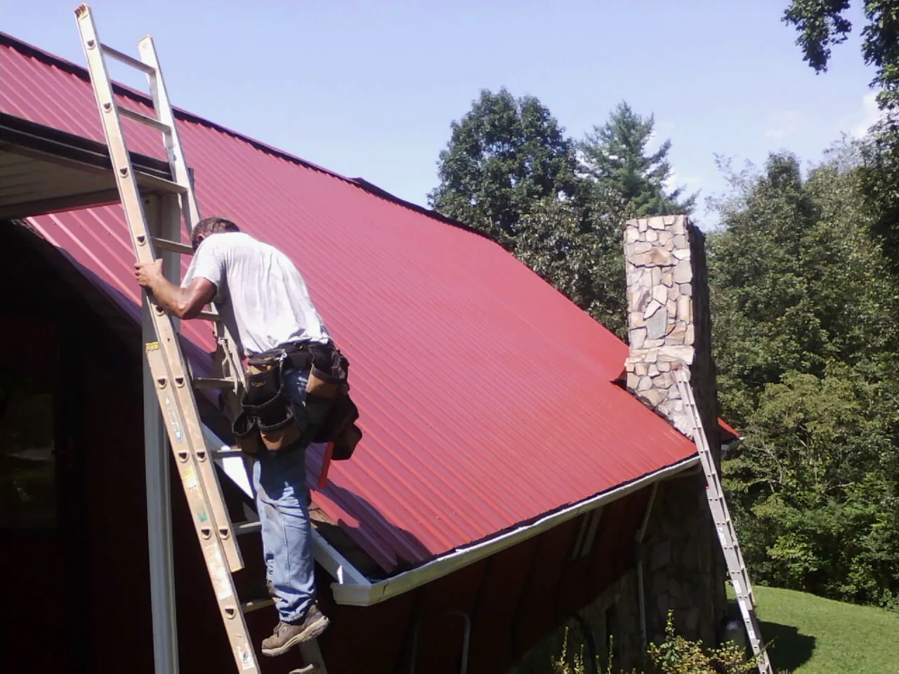 Roofer on a ladder beside a red metal roof and stone chimney — Soffit & Fascia Repair by Roofing Brothers in Simms